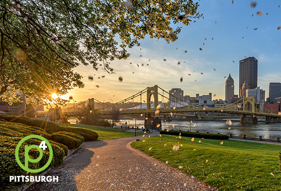 A walking path along a river with the sun setting in the distance. A city skyline on the other side of the river.